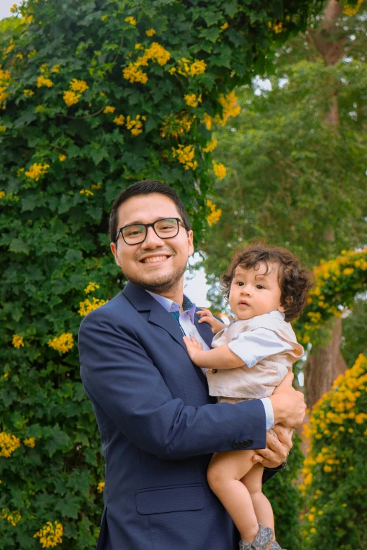 a man in glasses holding a baby next to green bushes