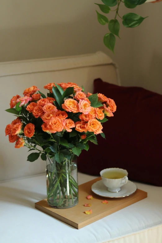 a vase of orange roses with teacup and saucer sitting on a bed