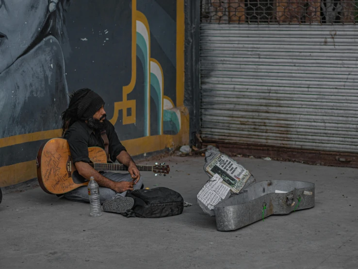 a man with dreadlocks is sitting on the sidewalk playing guitar