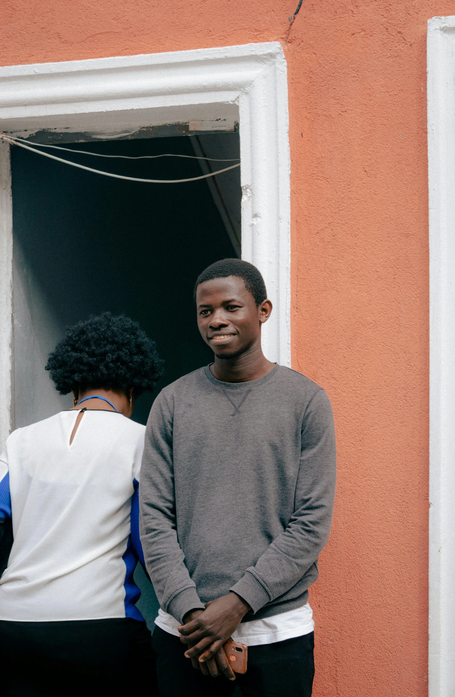 two men stand outside of a small building, with one smiling