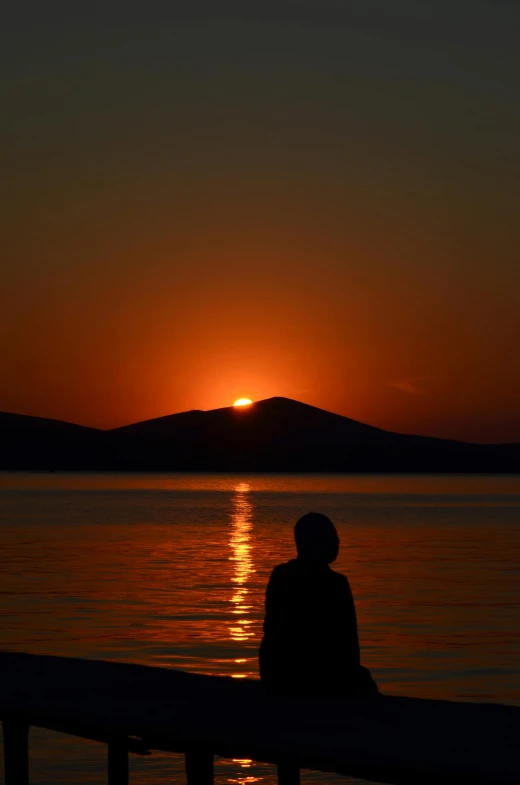 silhouette of person sitting on pier with setting sun