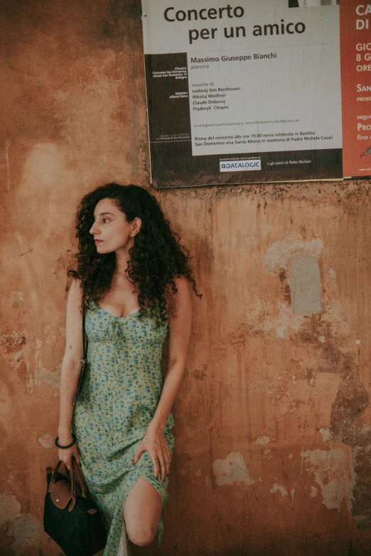 a woman in a floral dress stands against a wall with her bag in hand