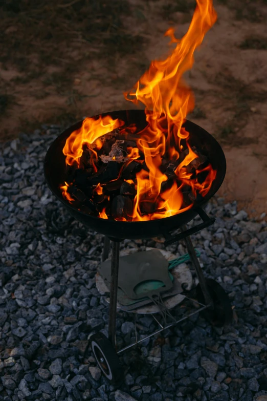 a charcoal grill with orange flame is on gravel next to a fire pit