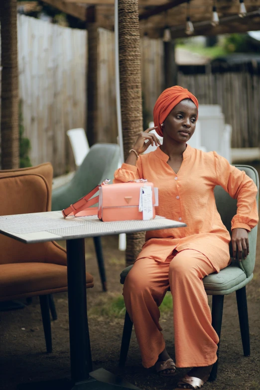 a black woman in orange is sitting at a table with her hand on her telephone