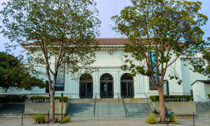 the front entrance of a white building with a staircase and trees in front