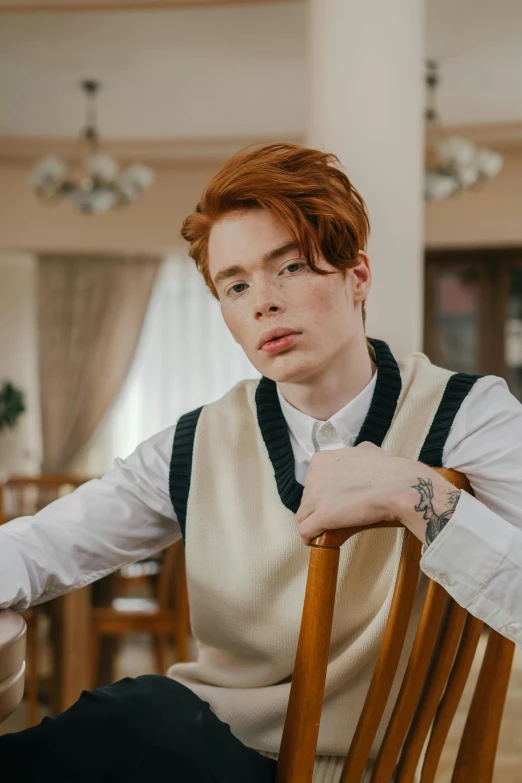 a young man with red hair wearing a sweater and a vest is seated in a chair