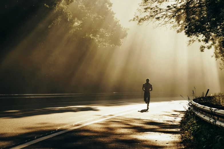 the man is jogging down the street while the sun is shining through the trees