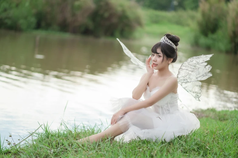 an asian girl dressed in white and holding a feather sits on the grass by water