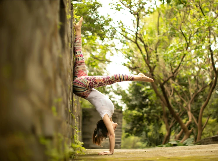 a  standing on one arm and leaning over with the hand on the wall and her leg up in the air, against a stone wall near a brick wall