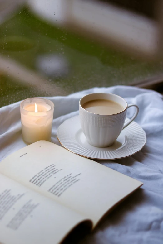 an open book and a cup of coffee sit on a bed next to a candle