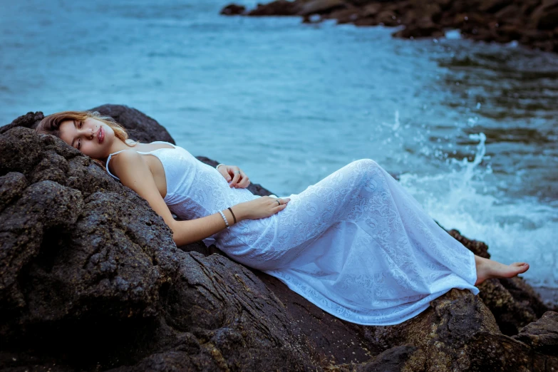 a pregnant woman laying on top of a rock by the ocean