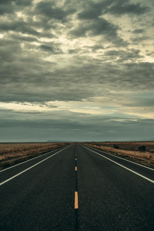 a long empty road is surrounded by brown hills and a cloudy sky