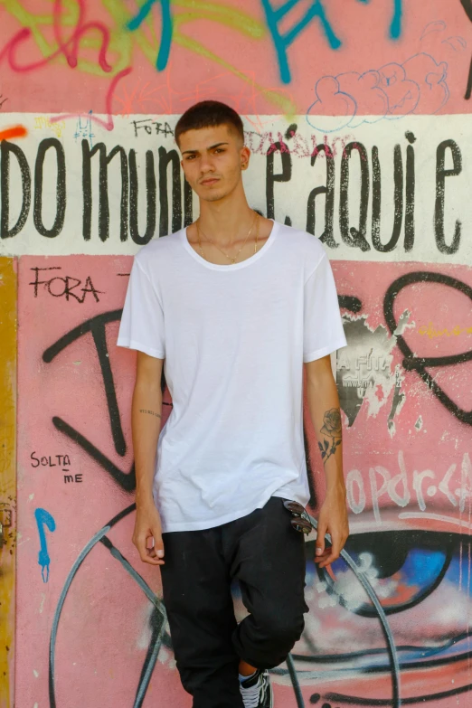 a guy wearing black pants, a white t shirt, and converse shoes is standing in front of a wall with graffiti