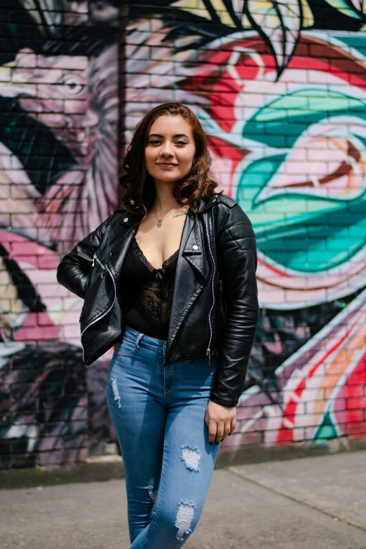 a woman wearing jeans and a black jacket standing in front of a graffiti wall