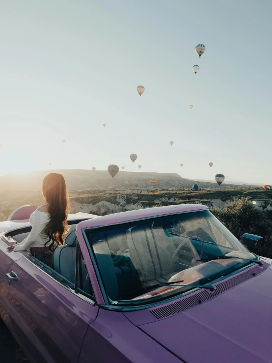 a woman sitting in a convertible car with  air balloons flying above