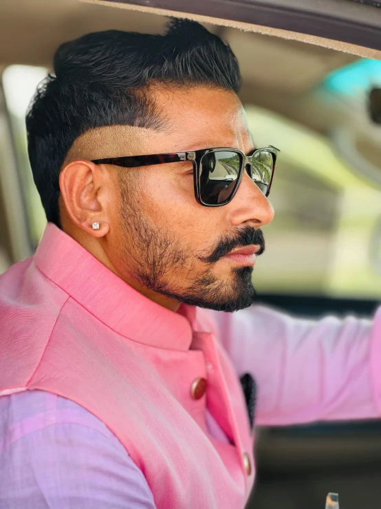 a man in sunglasses drives a car with a pink shirt on