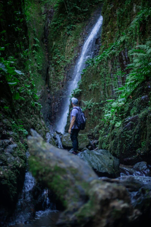 man walking on rocks towards large waterfall in dense tropical forest