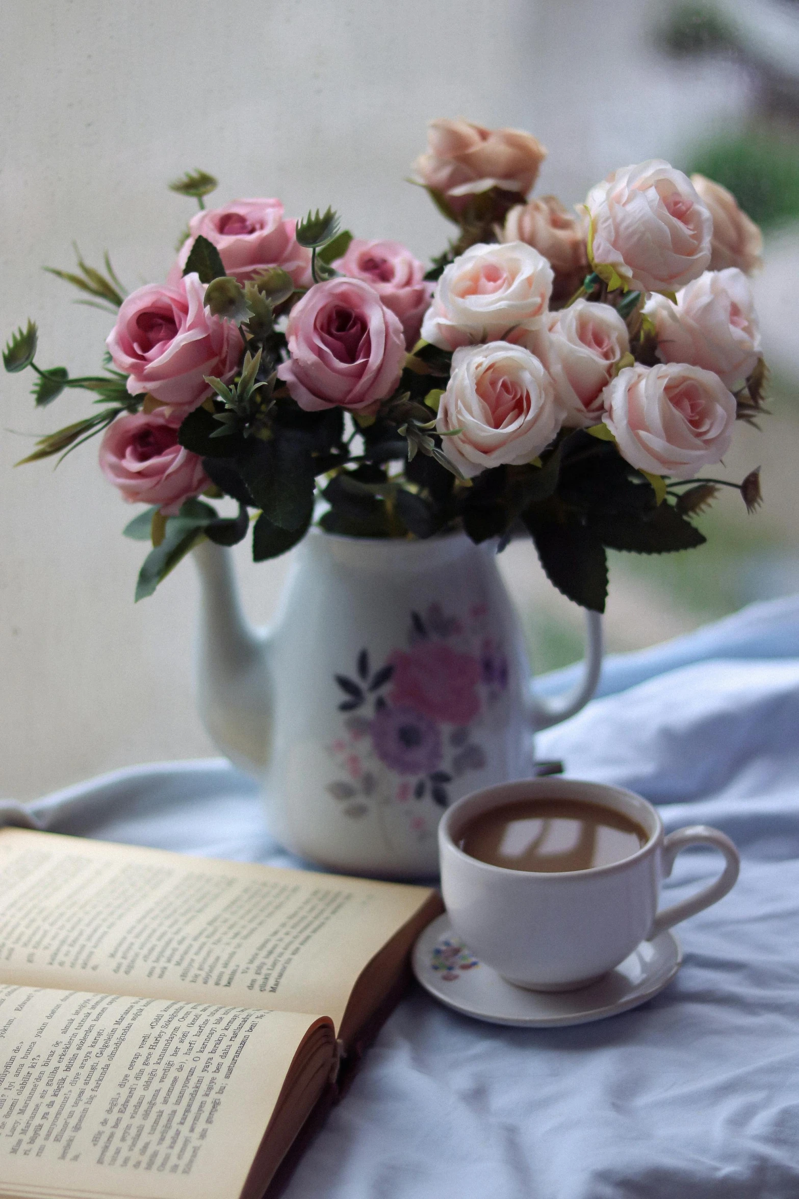 a cup of coffee and pink flowers sit next to an open book on a bed