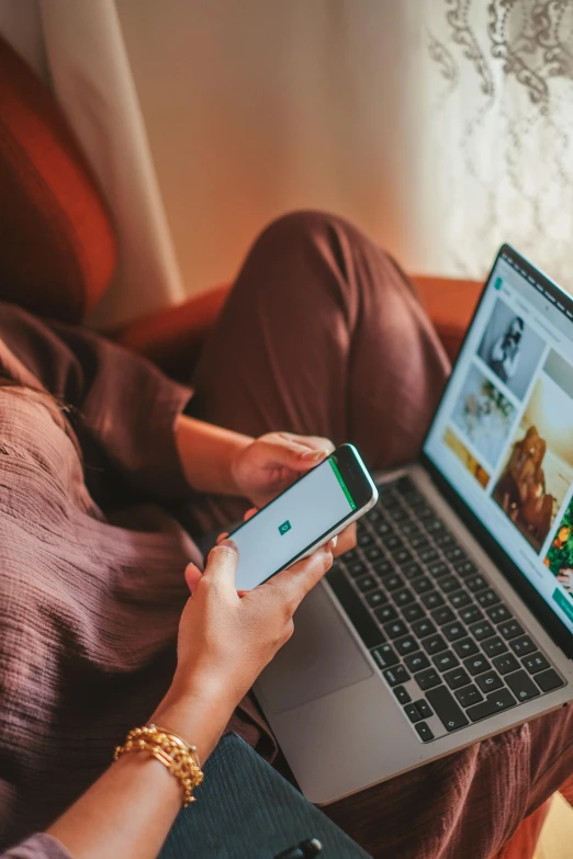 a woman in a brown sweater sits in a chair using a laptop with a cell phone and a green iphone