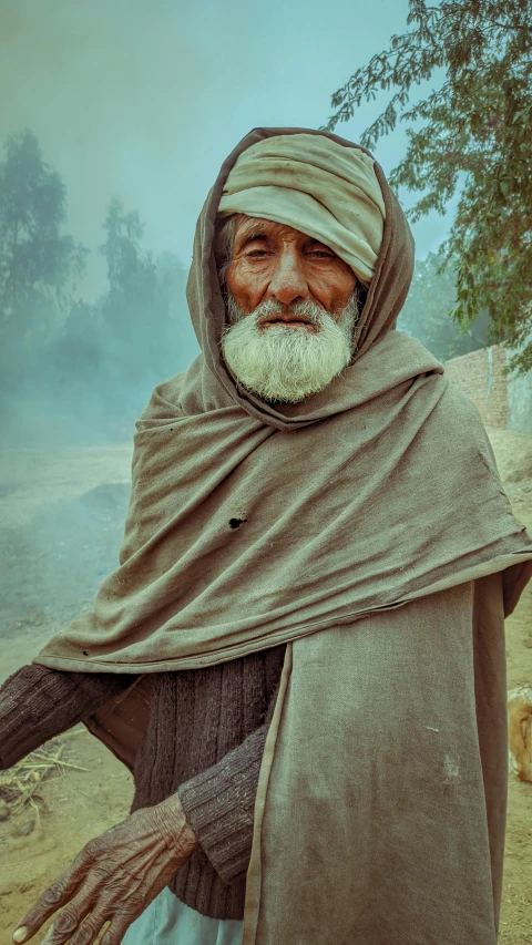 a man wearing a tan shawl standing in front of some trees