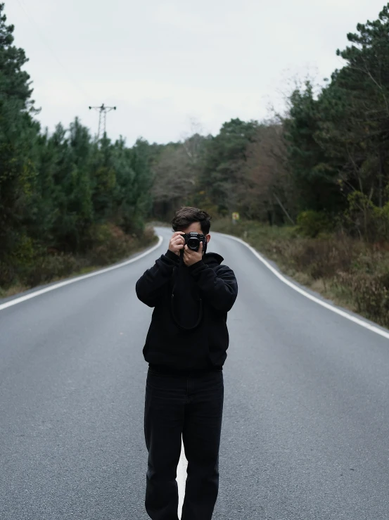 man wearing glasses standing in the middle of the road while holding camera up to his eye