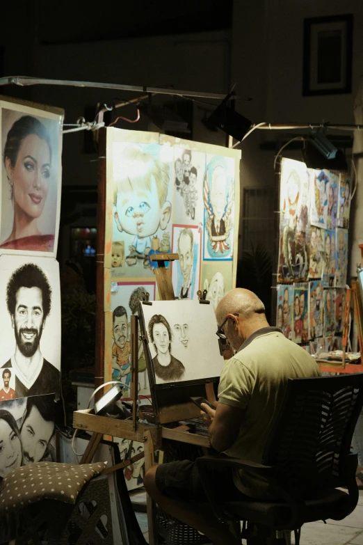 a man sits in a chair at an easel in front of posters on easels, with paintings and pos of the people in them