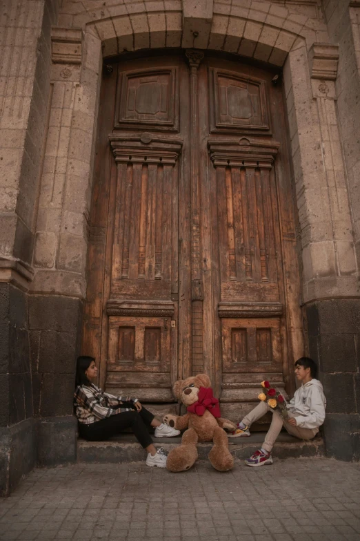 two people sitting on a curb with a teddy bear near an old building