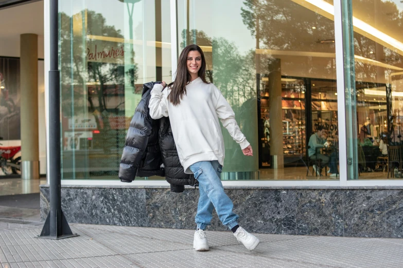 a woman in white coat and blue jeans walking on sidewalk