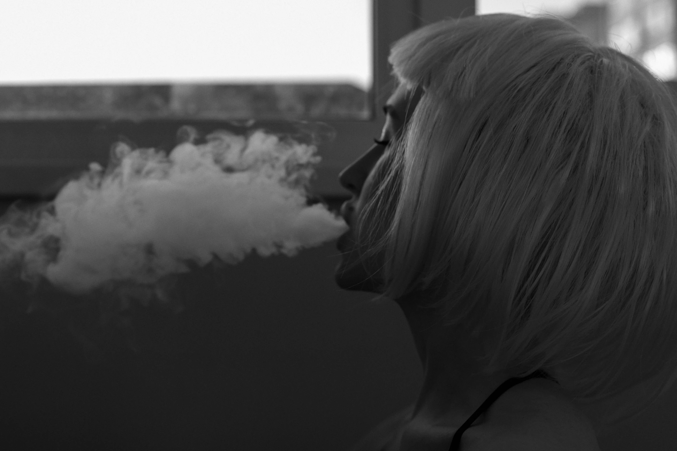 a blonde woman sitting at a window smoking a cigarette