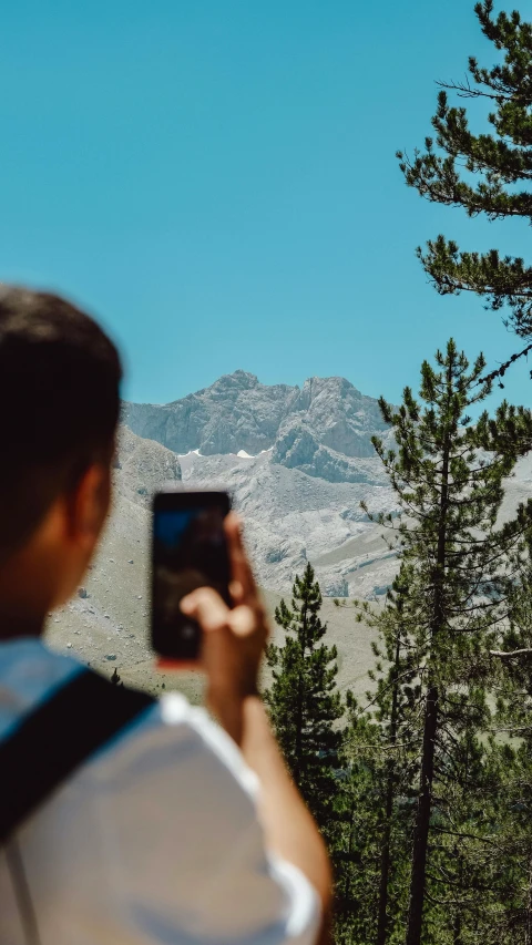 a man in white shirt and black backpack taking a po with cell phone of mountains and pine trees