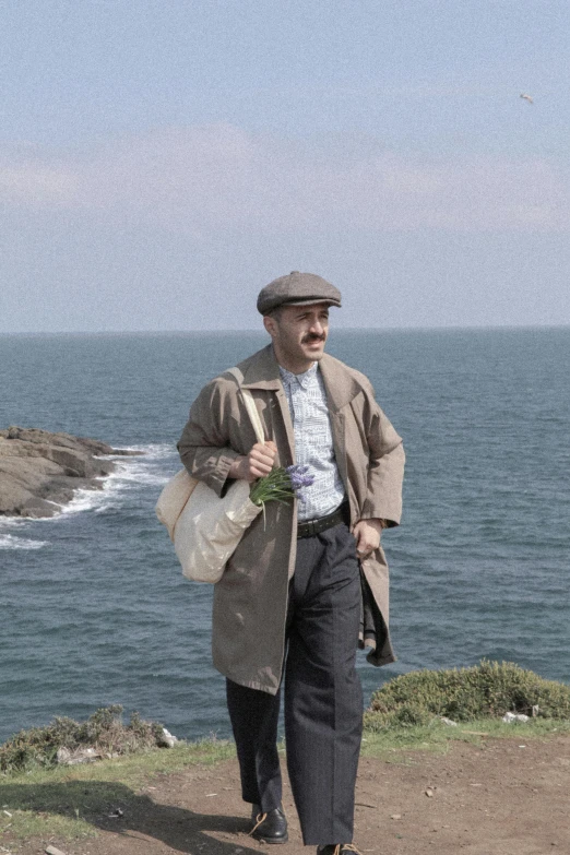 a man wearing a hat and coat walking by the ocean