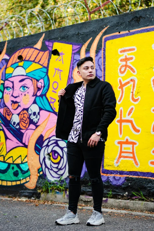 a young man stands next to a graffiti covered wall