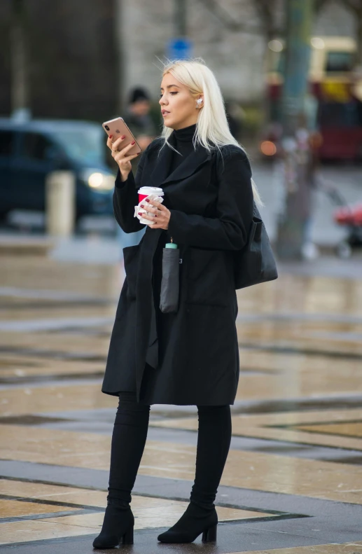 a woman in black jacket holding coffee looking at her phone