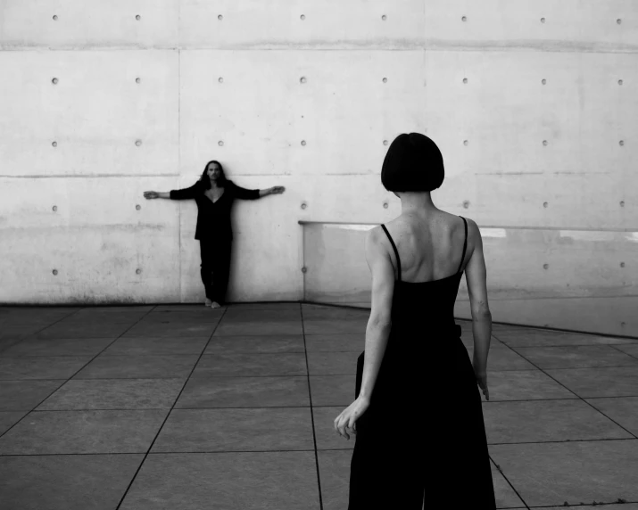 two women with black clothing stand by wall looking at one another