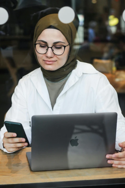 a girl sitting at a table holding a cell phone and looking at a laptop
