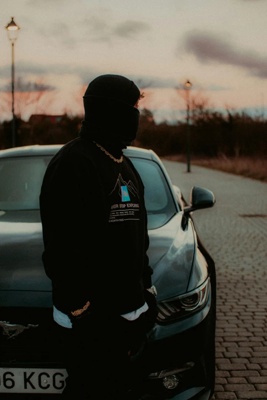 a young man stands in front of his car in the middle of a brick road