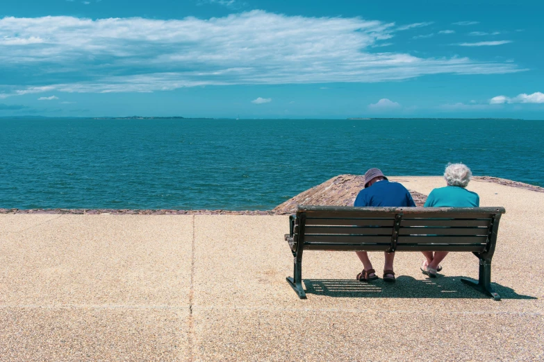 an older couple sits together on a bench looking out into the ocean