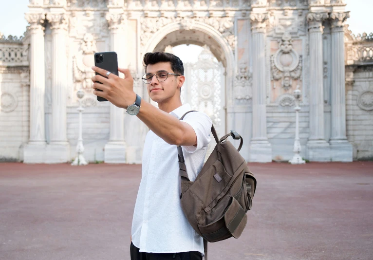 a young man wearing glasses, white shirt and a dark bag takes a selfie with his cell phone