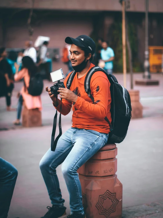 man in a red shirt, black hat, and backpack sits on the curb looking at his camera