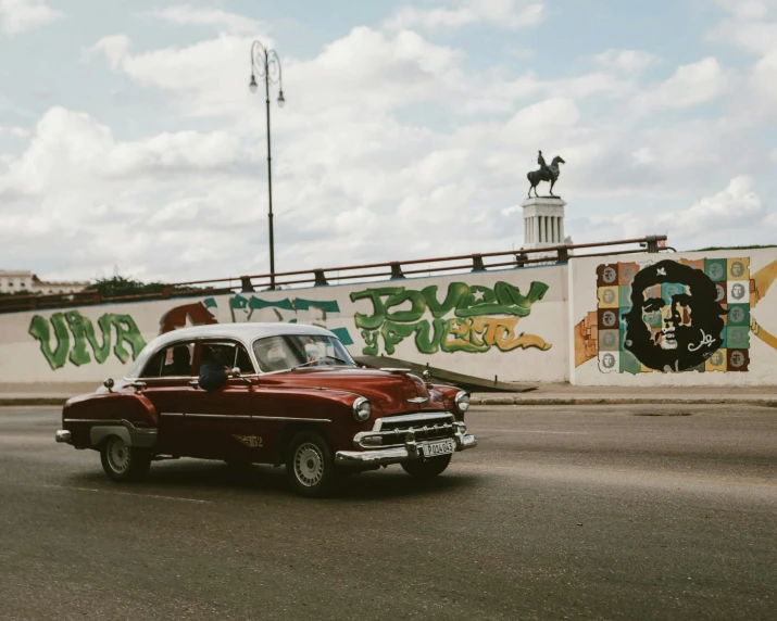 an old car passing by a large wall with colorful graffiti