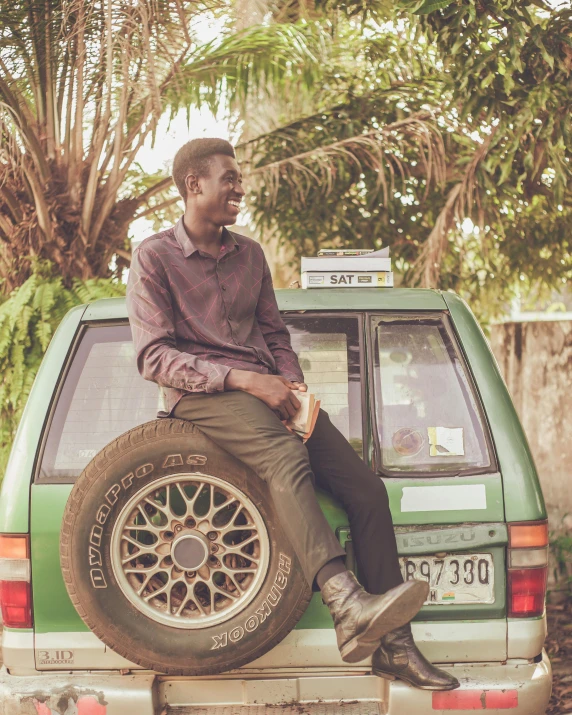 man sitting on tire of green car with green tree behind