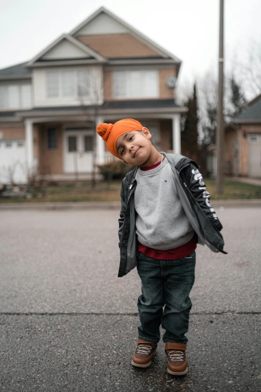 a child in an orange knit cap and a jacket stands on asphalt in front of two houses