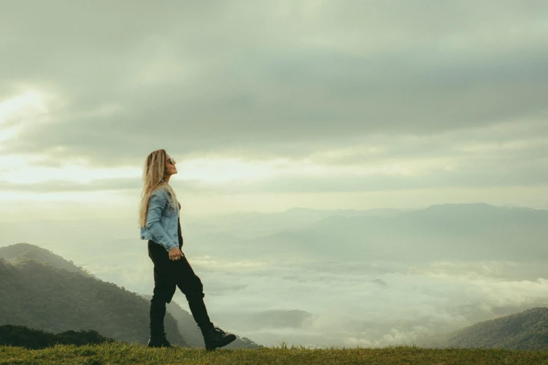 a young woman standing alone on a grassy hill looking up at the clouds