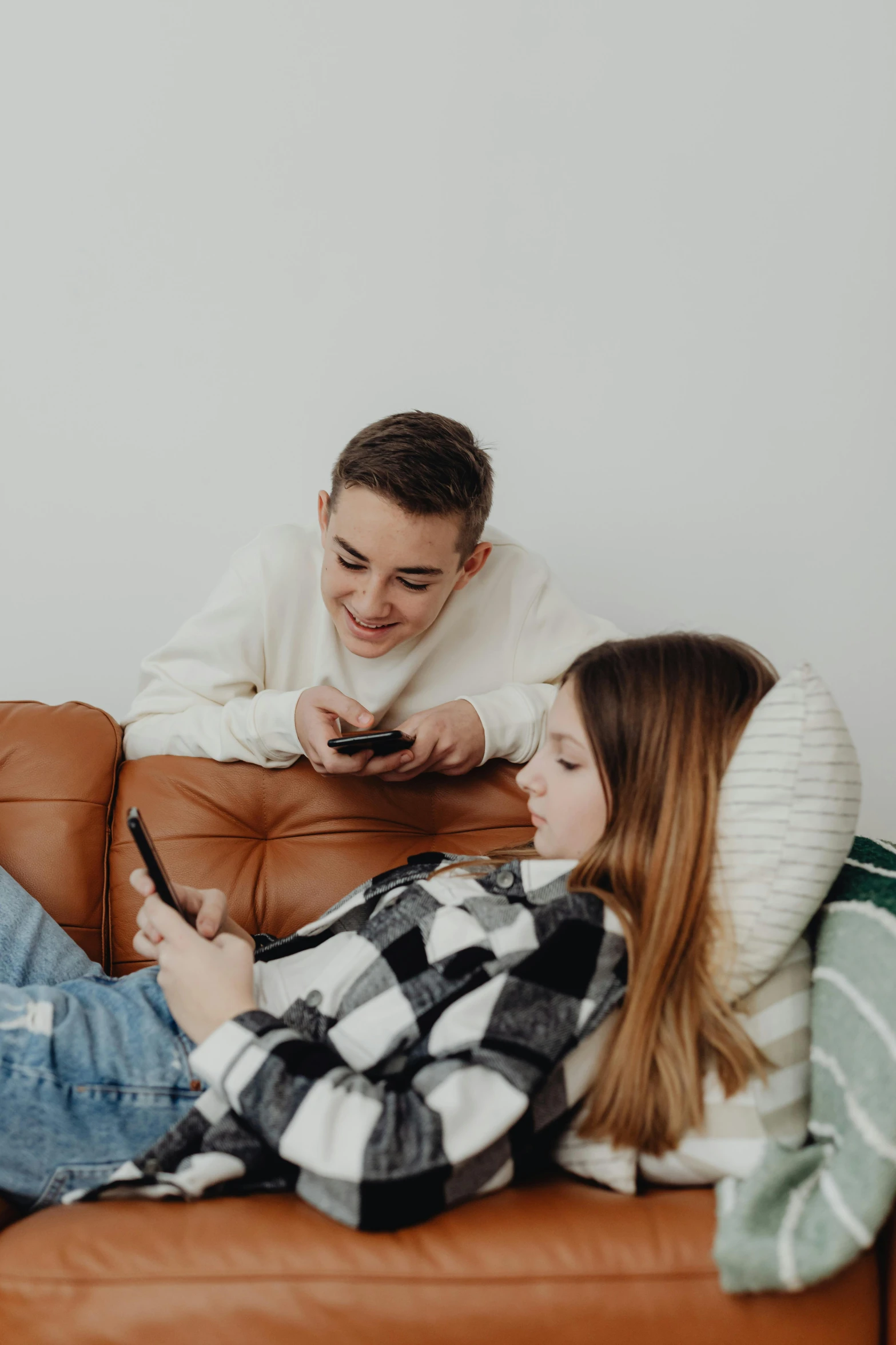 a couple sit on a brown leather couch and look at their cell phones