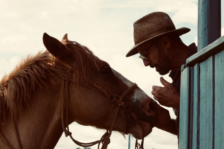 a man in straw hat leans on the side of a building with his horse