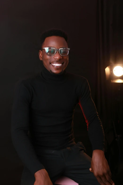 a black male sitting on a stool wearing a black sweater