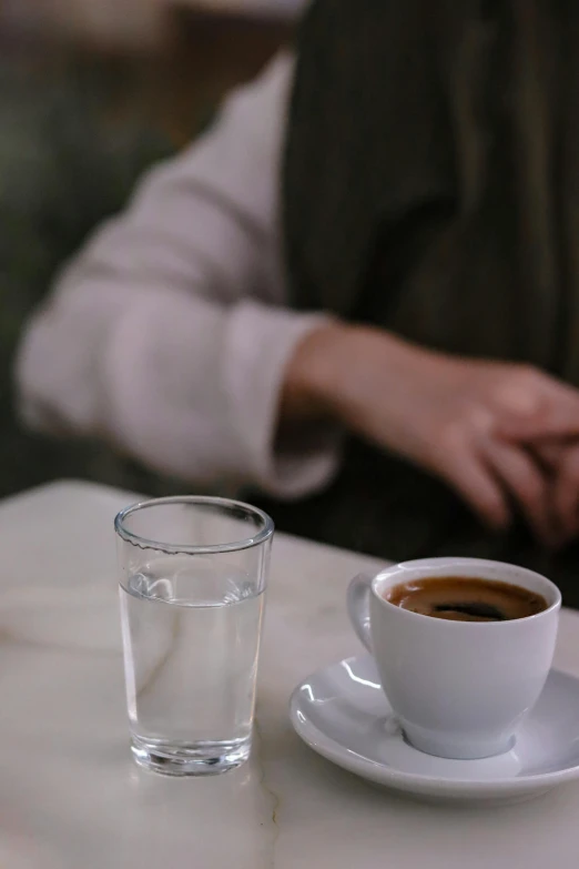 a glass on a white table in front of a cup of coffee