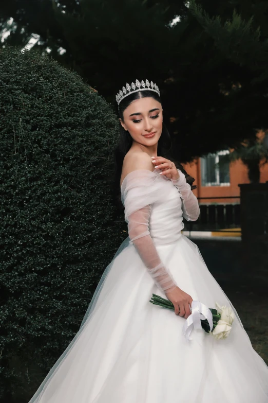 a girl in a white dress and a tiara is standing next to a tree