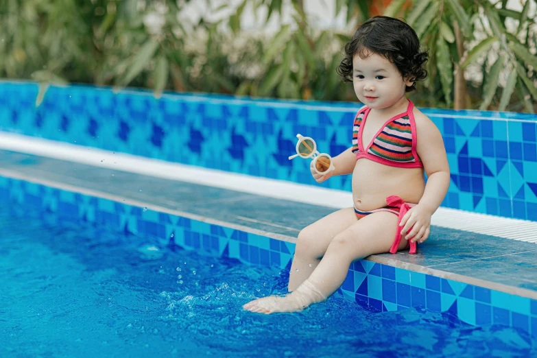 a little girl in a bikini eating some cake while sitting on a ledge near a pool