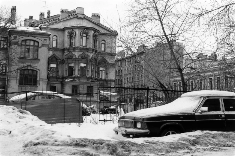 an old, abandoned, black and white car parked in front of a large mansion on a snowy day